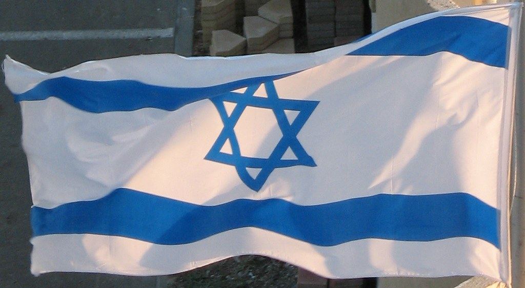 An Israeli flag with a blue Star of David and two blue stripes waving outdoors in sunlight