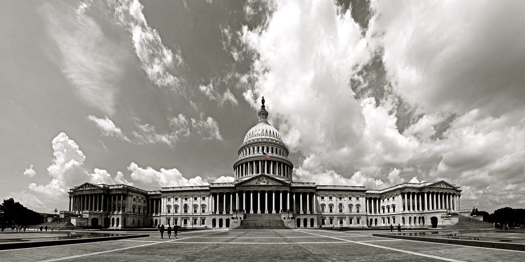 Black-and-white view of the U.S. Capitol building under dramatic clouds, with a wide plaza in front