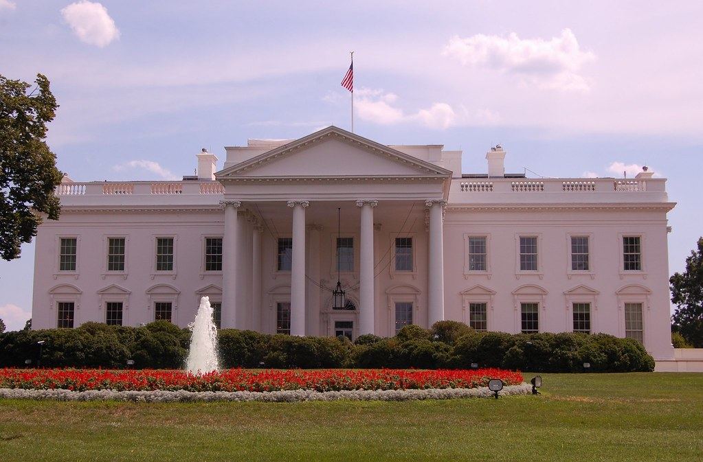 The White House north facade with an American flag, a fountain and red flower beds on the front lawn
