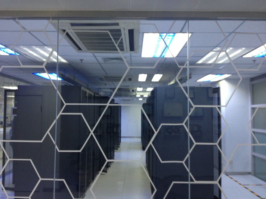 Rows of dark server cabinets in a brightly lit data center, seen through glass with a white hexagon pattern
