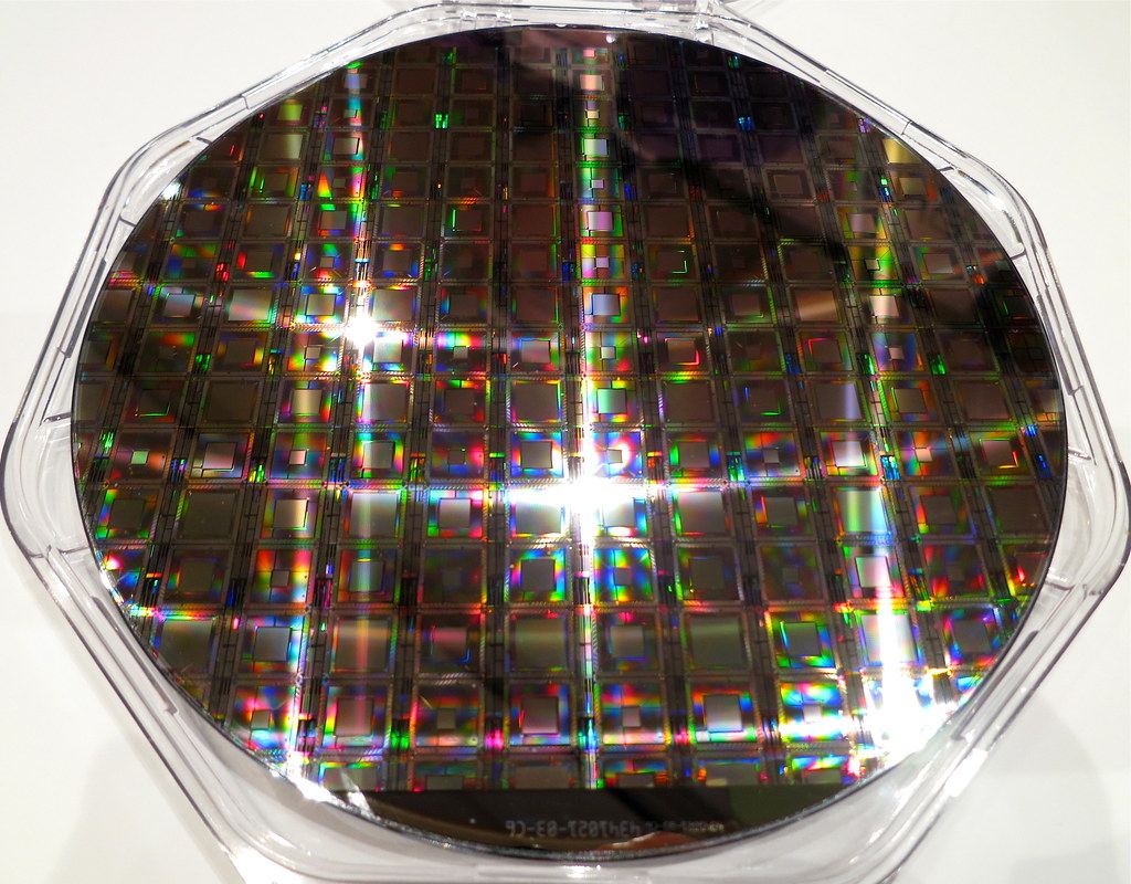 A silicon wafer covered in a grid of processor chips reflects rainbow colors inside a clear plastic case