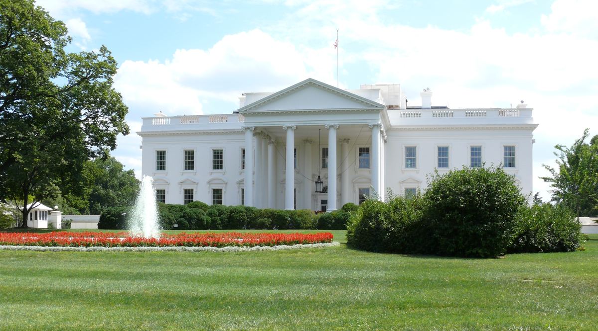 The White House north facade with a fountain, red flower beds and green lawn under a partly cloudy sky