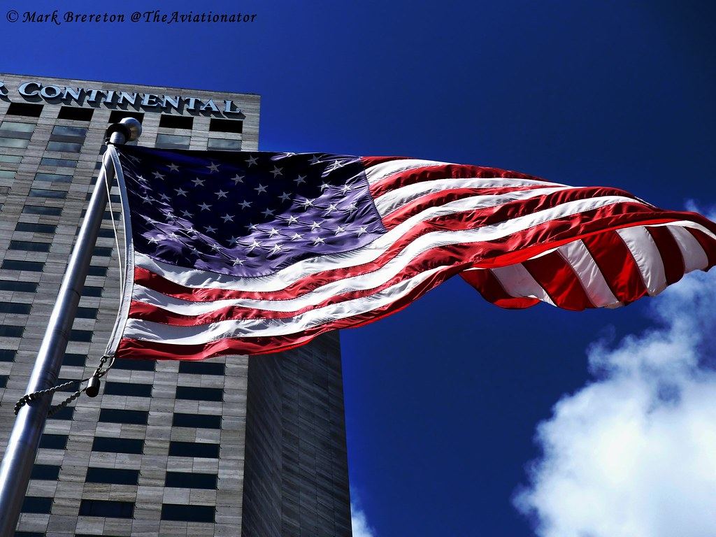 An American flag waves on a flagpole in front of a tall office building under a blue sky with clouds