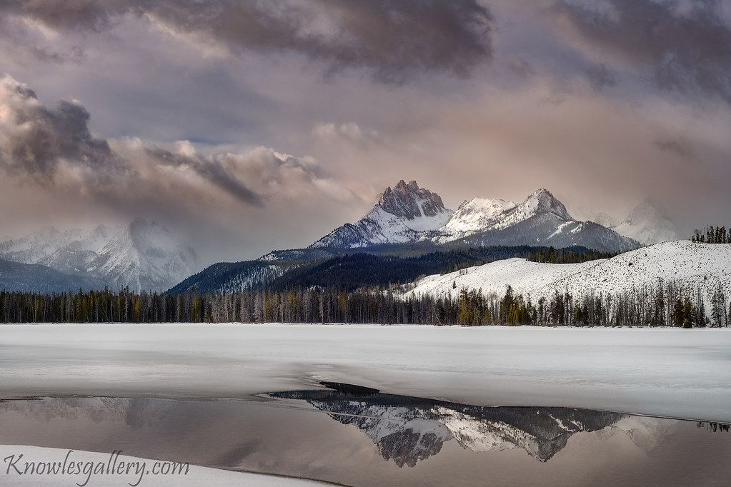 Snow-covered mountain peaks under pastel morning clouds, reflected in a partly frozen lake lined with pine trees