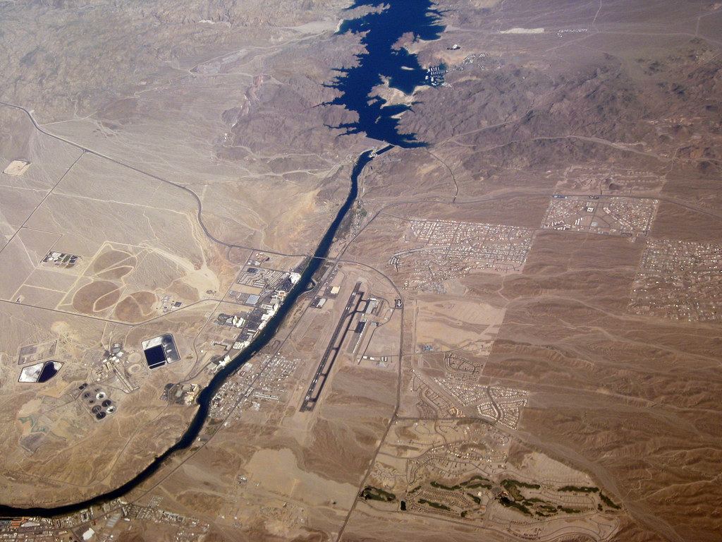Aerial view of a desert river below a dam and lake, with casinos, towns and an airport runway along its banks