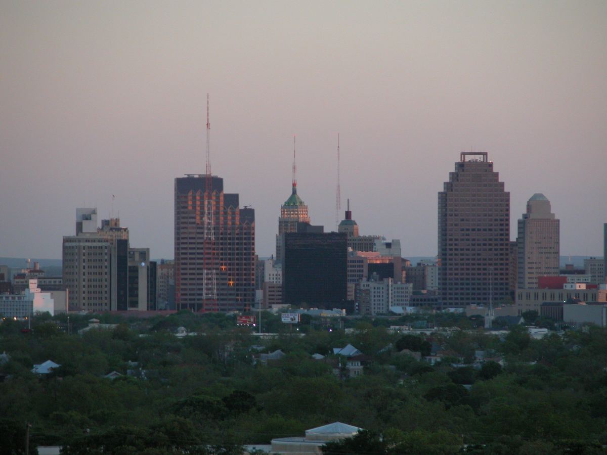 Downtown San Antonio skyline at dusk, seen from the north above a dense canopy of trees