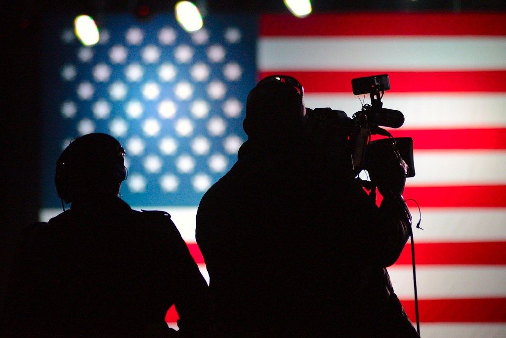 Silhouettes of two people, one holding a video camera, in front of a large illuminated American flag