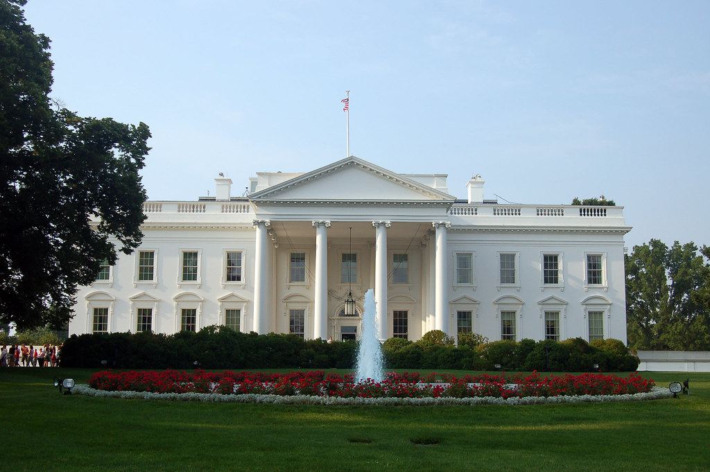 The White House north facade with columns, a fountain, red flower beds and a green lawn under a clear sky