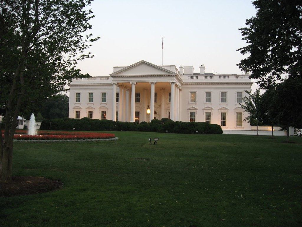 The north facade of the White House with white columns, a green lawn and a fountain at dusk