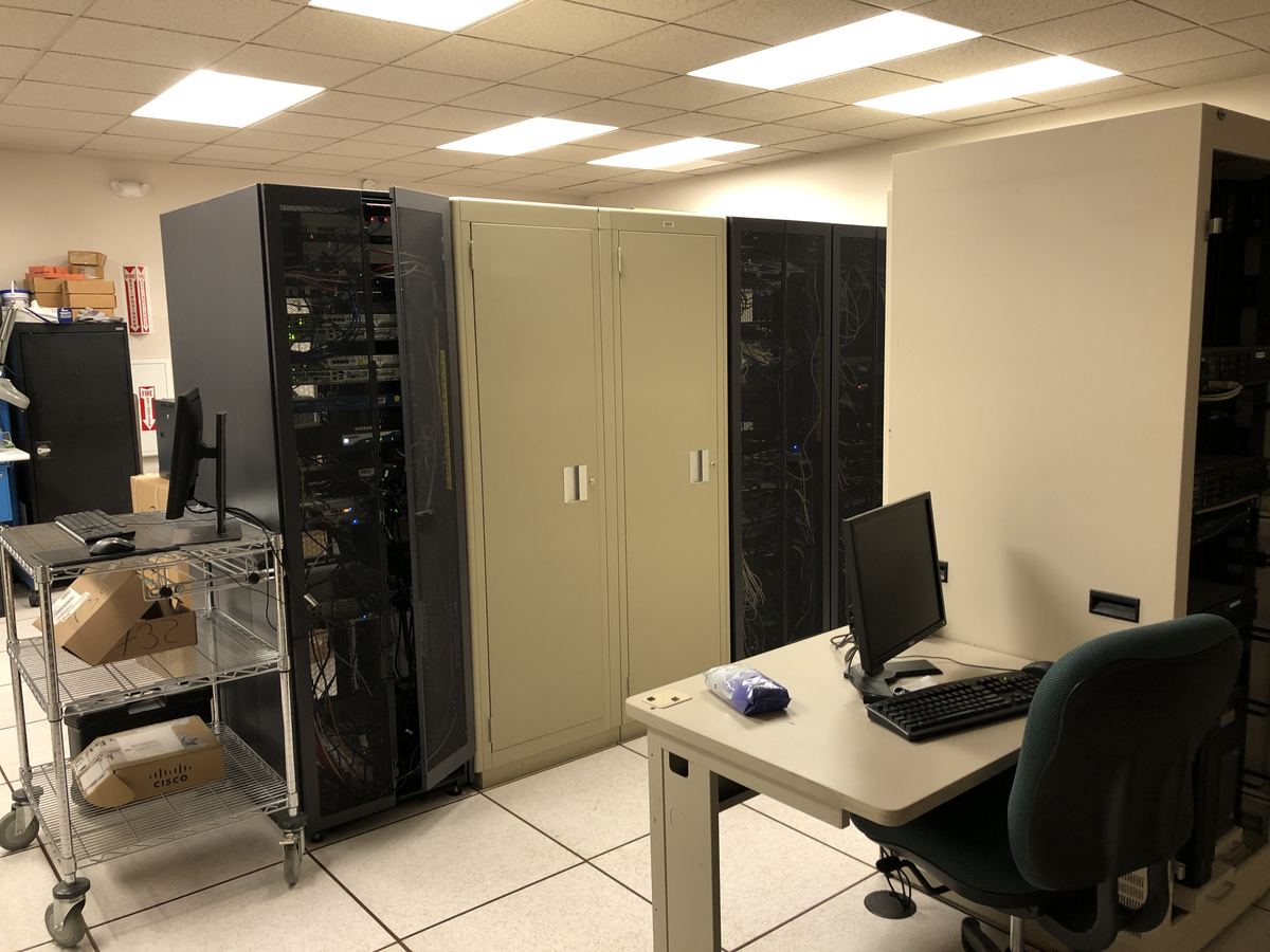 Server racks with tangled network cables beside beige cabinets, a computer desk and an equipment cart in a tiled room