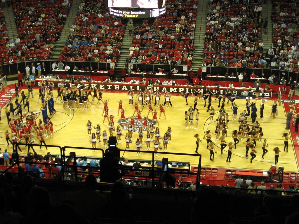 Cheerleading squads perform on a basketball court inside a crowded arena with red seats