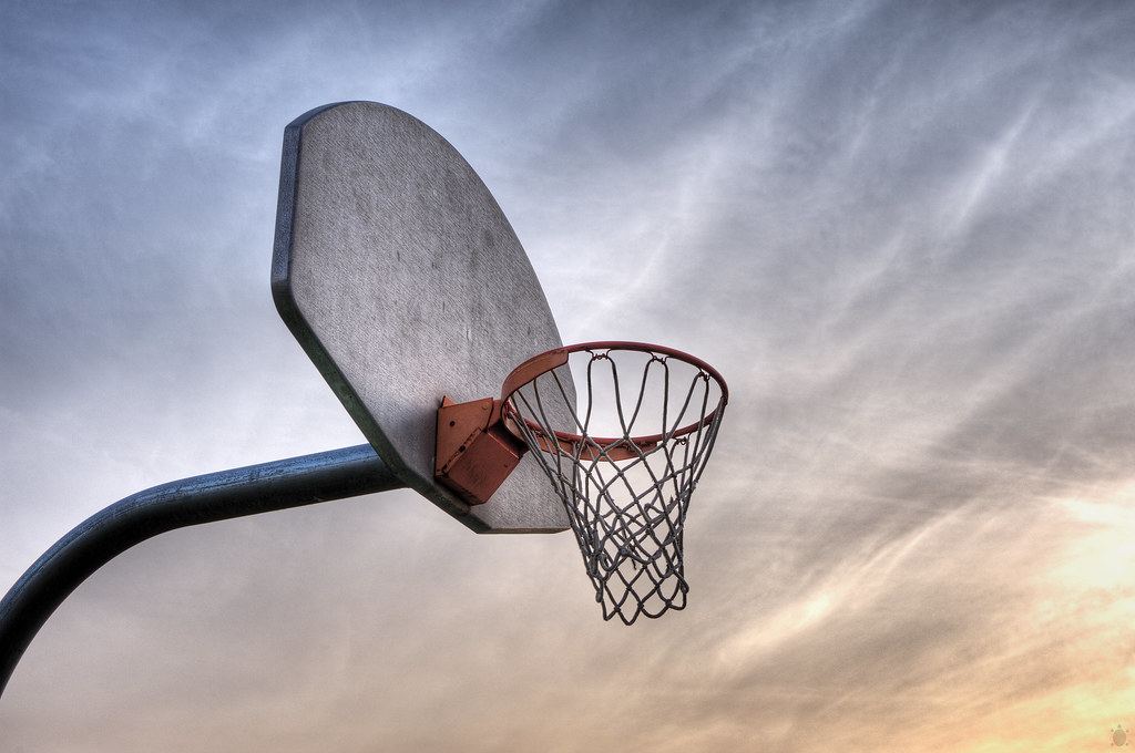 An outdoor basketball hoop with a metal backboard and chain-style net against a cloudy sky at sunset