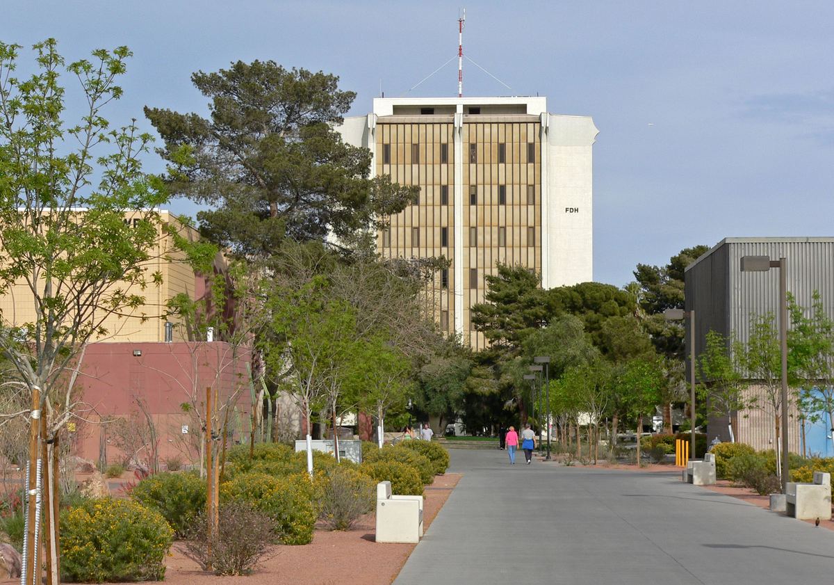 A tree-lined campus walkway leads toward a tall tan building marked FDH under a blue sky, with desert landscaping
