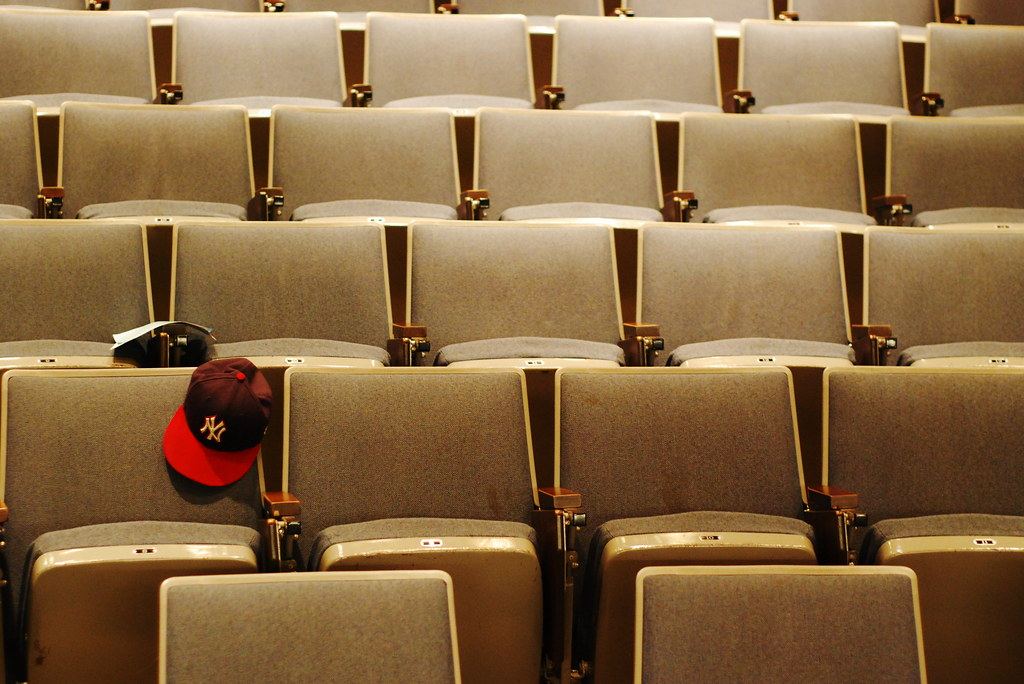 Rows of empty gray upholstered seats in a lecture hall, with a red and maroon baseball cap hanging on one seat