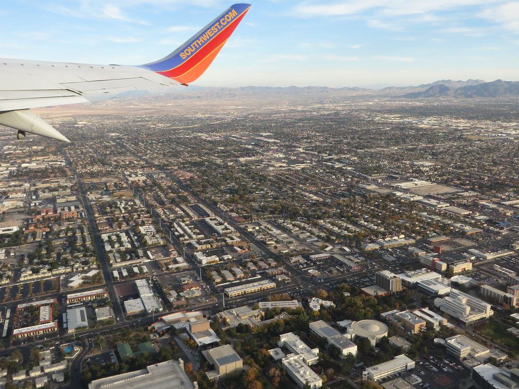 Aerial view from an airplane window of a sprawling city grid with campus buildings in front and mountains beyond