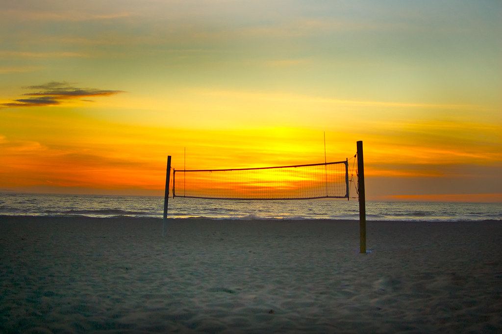 A volleyball net stands on an empty sandy beach in front of the ocean under an orange sunset sky