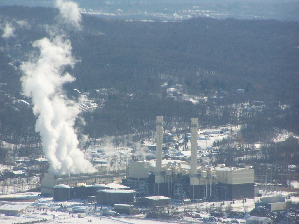 A power plant with two tall stacks and a large steam plume beside snowy, wooded hills