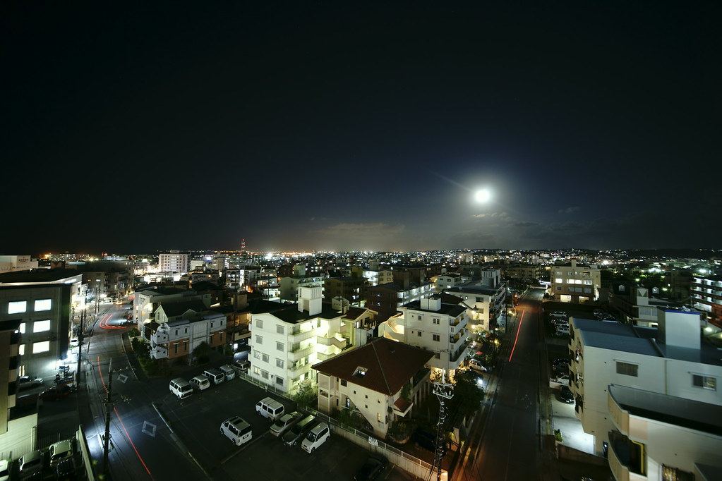 Night view over low-rise apartment buildings, lit streets and parked cars, with a bright moon above the city