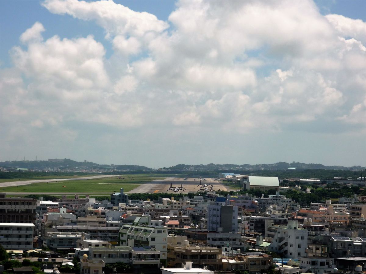 Dense city buildings in front of an airfield runway with tiltrotor aircraft under a cloudy sky