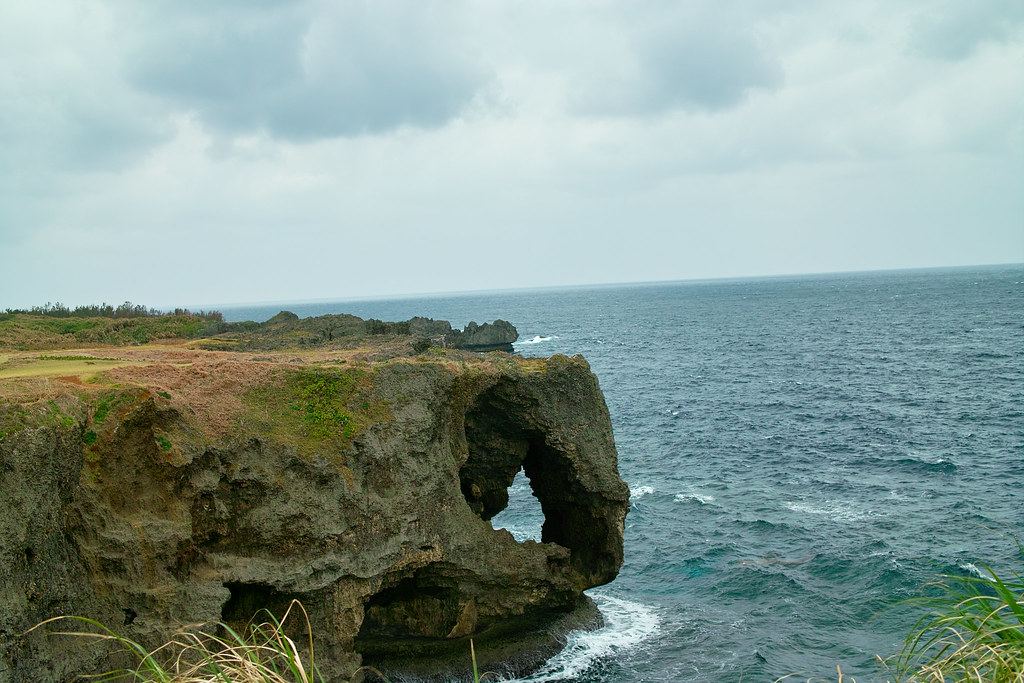 A rocky limestone cliff with a natural arch juts into the dark blue sea under a cloudy sky