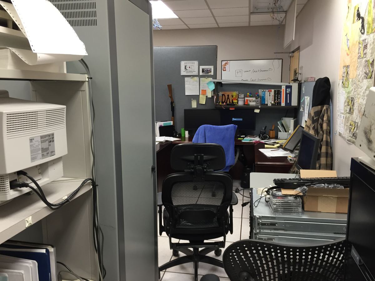 A cluttered office workspace with desk chairs, computer monitors, a printer and rack-mounted server equipment