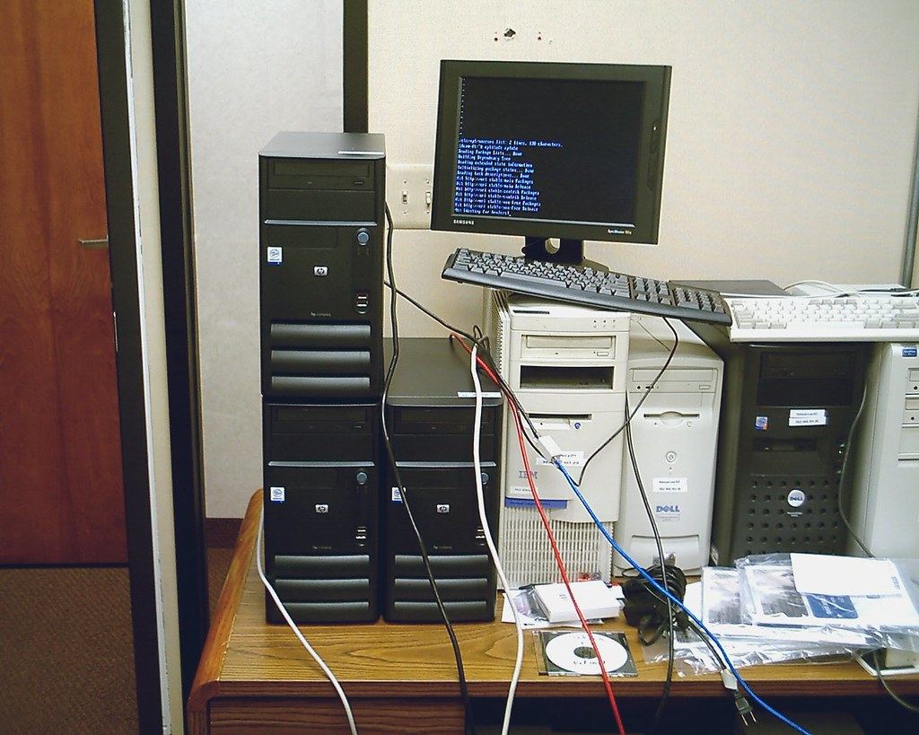 Several desktop computer towers, a monitor showing text and a keyboard crowded on a wooden desk with tangled cables