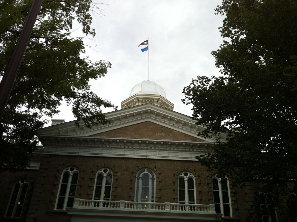 Sandstone capitol building with a silver dome and US and Nevada flags, framed by trees under a cloudy sky