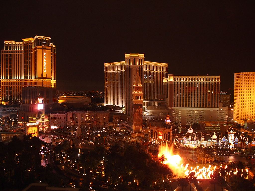 Las Vegas Strip hotel towers lit up at night, with palm trees and a fiery fountain show in the foreground