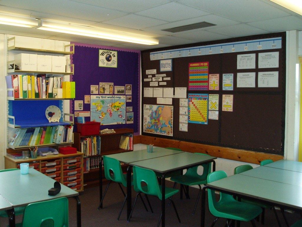 An empty classroom with green chairs, grey tables, bookshelves and maths and world map posters on the walls